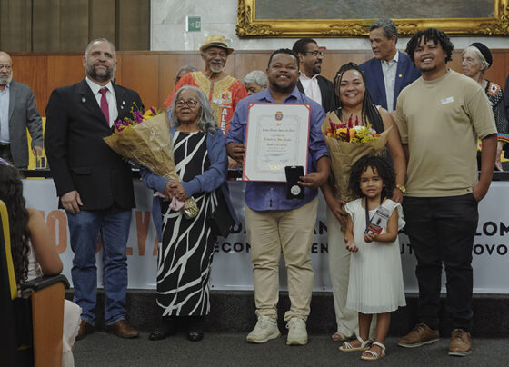 Foto de grupo com sete pessoas em pé. Homem ao centro segura diploma e medalha; duas mulheres com buquês; criança à frente. Ao fundo, mesa com autoridades e moldura dourada. Ambiente interno, iluminação clara e clima cerimonial.