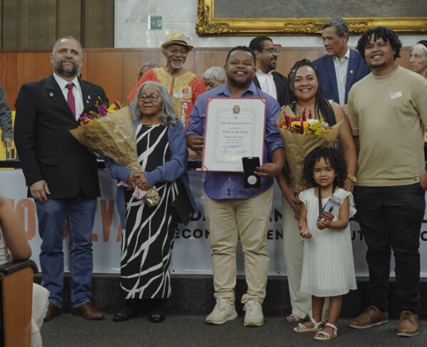 Foto de grupo com sete pessoas em pé. Homem ao centro segura diploma e medalha; duas mulheres com buquês; criança à frente. Ao fundo, mesa com autoridades e moldura dourada. Ambiente interno, iluminação clara e clima cerimonial.