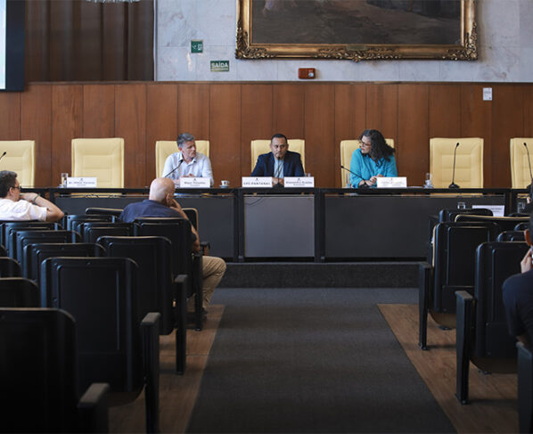Imagem de uma mesa de audiência com três pessoas sentadas e microfones. Placas de identificação à frente. Auditório com público pequeno, cadeiras pretas, parede de madeira e quadro grande ao fundo. Luz natural suave.