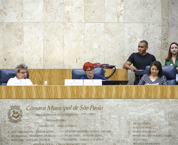 Imagem de sessão na Câmara Municipal de São Paulo. Três pessoas sentadas à mesa e três em pé ao fundo. Parede de mármore clara com brasão e inscrições gravadas. Iluminação forte e ambiente formal.