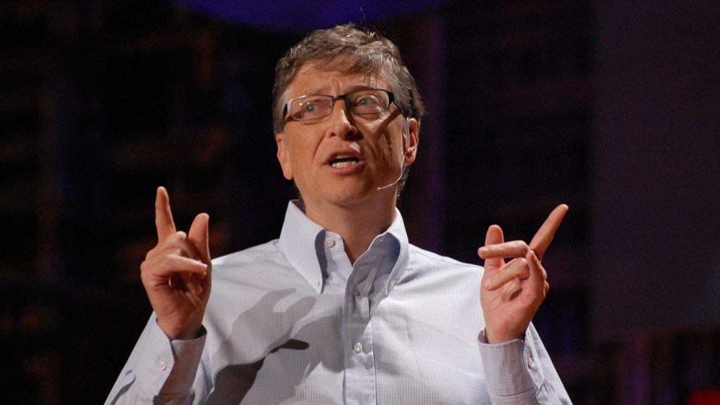Bill Gates em palestra, apontando com as mãos para cima. O fundo da imagem é preto