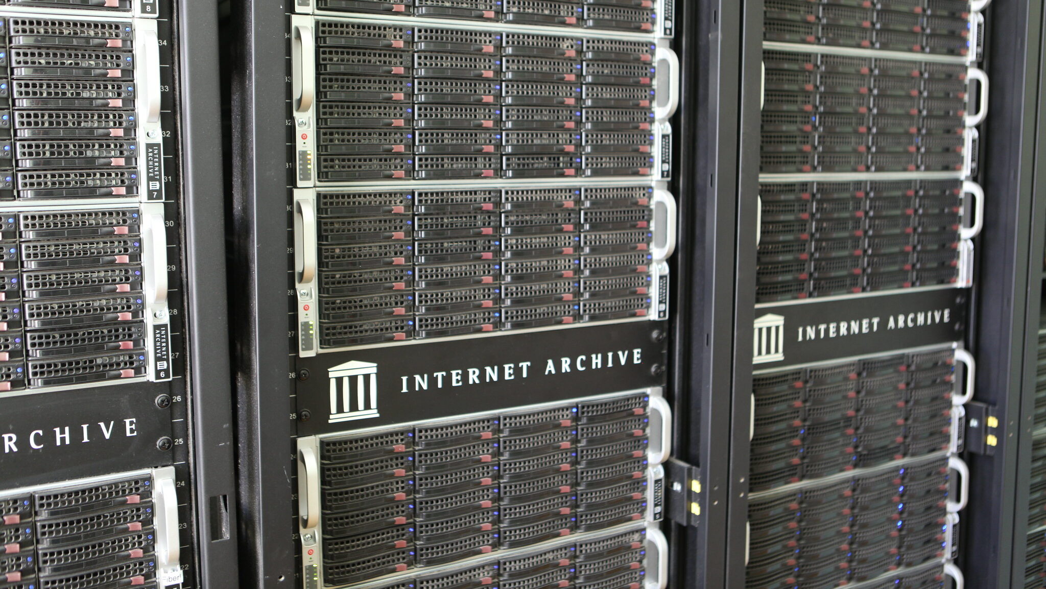 Fotografia em plano médio e ângulo diagonal mostra três racks de servidores pretos alinhados lateralmente. Os equipamentos possuem diversas gavetas horizontais com frentes gradeadas e pequenas luzes indicadoras acesas em tons de azul e vermelho. No centro de cada rack, há uma placa preta com o logotipo de um templo grego e o texto "INTERNET ARCHIVE" em letras brancas e maiúsculas. Nas laterais, veem-se etiquetas brancas numeradas e alças de metal prateadas para manuseio.