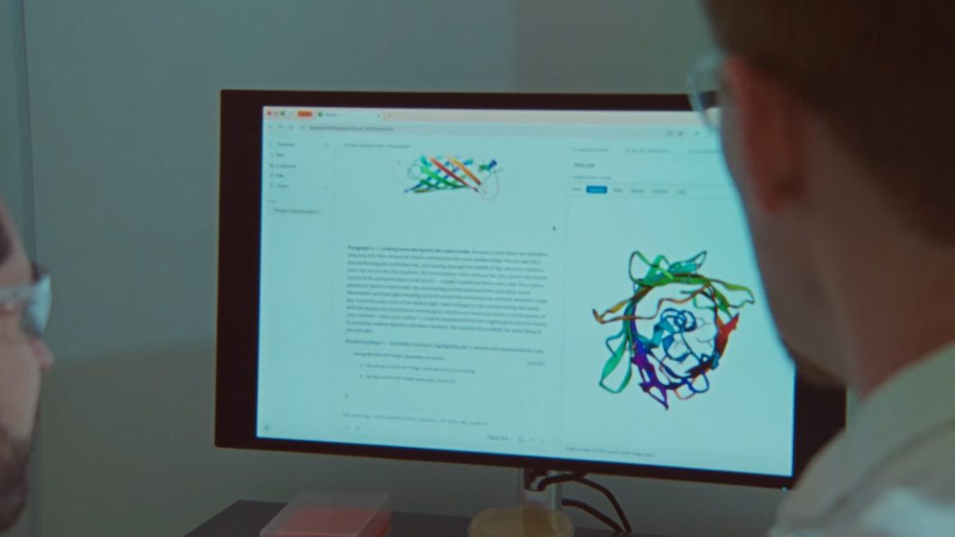 Fotografia de um pesquisador observado de costas à direita enquanto analisa um monitor de computador; a tela exibe uma interface de software científico com texto explicativo e duas representações tridimensionais de estruturas moleculares de proteínas em fita colorida.