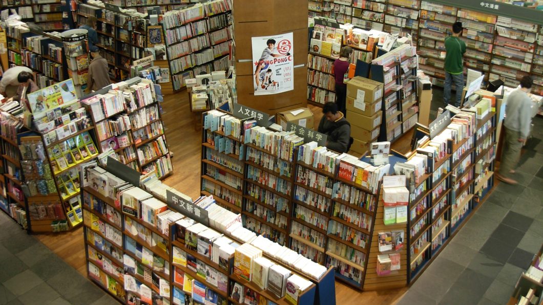 Fotografia em vista aérea do interior movimentado de uma livraria japonesa de grande porte; o ambiente é repleto de altas estantes de madeira organizadas em corredores repletos de livros e revistas com placas indicativas em ideogramas orientais (Kanji/Kana), enquanto clientes e funcionários circulam ou examinam os produtos entre as seções.